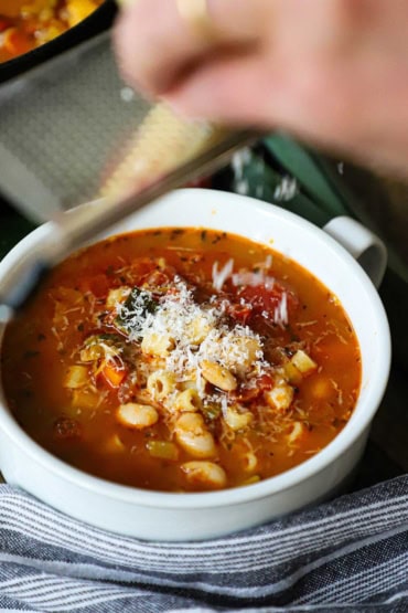 A straight-on view of a person grating a block of Parmesan cheese across a microplane over a soup bowl filled with classic minestrone soup.