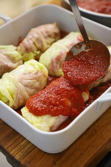 A person using a medium-sized ladle to pour on a tomato sauce over stuffed cabbage rolls in a white square baking dish.