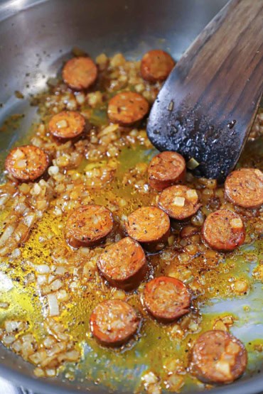 A straight-on view of a wooden spatula being used to stir and sauté chopped onions and slices of Spanish chorizo in a large stainless skillet with olive oil.