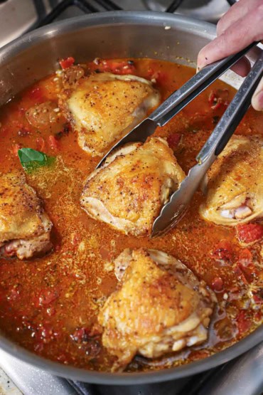 A person using a pair of metal tongs to turn a seared skin-on chicken thigh in a large skillet will with a simmering red sauce along with other seared chicken thighs.