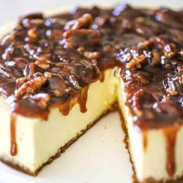 A close-up view of a cheesecake topped with a pecan pie sauce with a slice of cake missing.