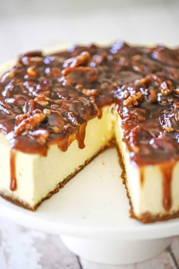 A close-up view of a cheesecake topped with a pecan pie sauce with a slice of cake missing.
