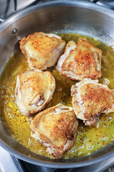 A straight-on view of five skin-on, bone-in chicken thighs that have been seared in a large skillet in olive oil and the skin visible and is browned and crispy.