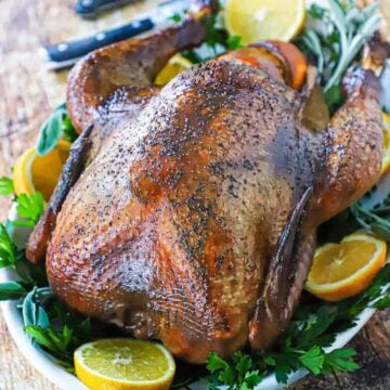 A straight-on view of a whole smoked-turkey that is resting on a white platter lined with fresh parsley and orange slices.