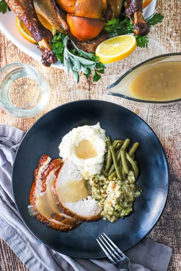 An overhead view of a black dinner plate filled with slices of smoked turkey topped with gravy and next to a mound of mashed potatoes, green beans, and dressing.