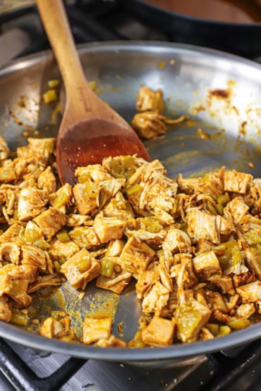A straight-on view of a large silver skillet that is filled with bite-sized pieces of leftover turkey in a red sauce and chopped green chiles being stirred with a wooden spatula.