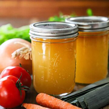 A straight-on view of two glass Mason jars that are filled with homemade vegetable stock with lids fastened on the tops of the jars and vegetables surrounding them, too.