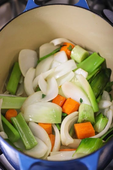 An overhead view of roughly cut onion, carrots, celery, and onion in an oval Dutch oven and are being sautéed.