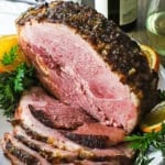 A straight-on view of a half smoked bourbon glazed ham resting on a large white platter with numerous slices cut from the roast on stacked also on the platter.