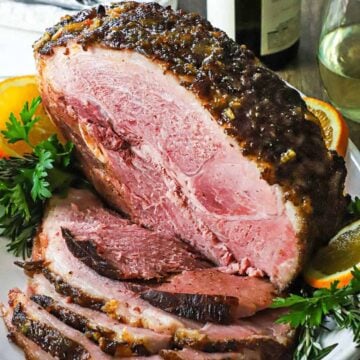 A straight-on view of a half smoked bourbon glazed ham resting on a large white platter with numerous slices cut from the roast on stacked also on the platter.