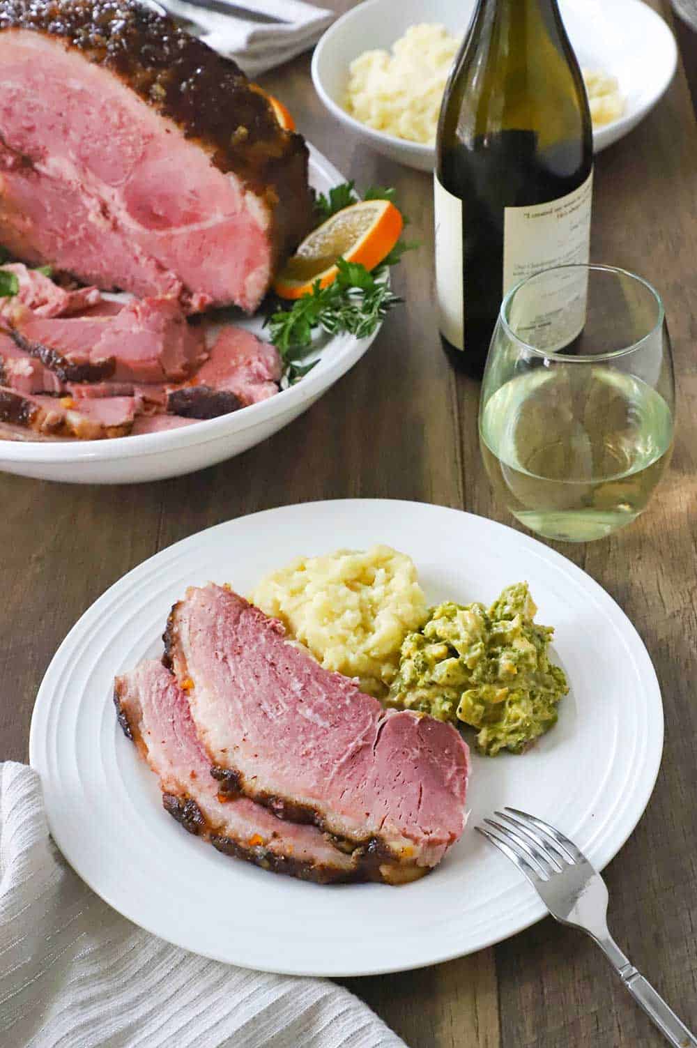 A white dinner plate with a serving of sliced smoked ham, mashed potatoe, and cheesy broccoli on it, with a glass of white wine and a platter of the sliced ham nearby.