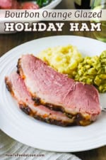 A close-up view of two thick slices of two thick slices of bourbon orange glazed ham on a white dinner plate with a serving of mashed potatoes and cheesy broccoli next to it.