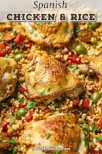 A close-up view of Spanish chicken and rice in a skillet with strips of roasted bell peppers and green olives scattered around the chicken in the rice.
