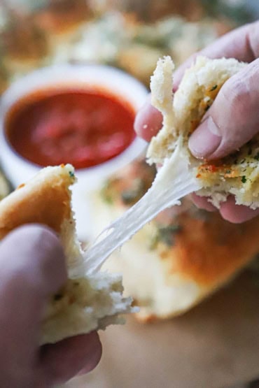 A person pulling apart a cheesy skillet rolls with two hands revealing a melty cheese pull with a bowl of marinara in the background. 