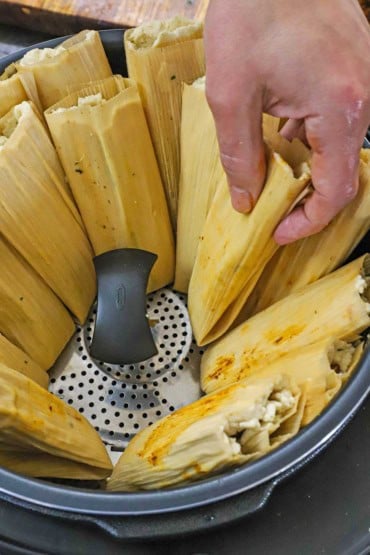 A person placing un-cooked homemade tamales into a pressure cooker with a steaming basket in the bottom.