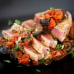 A close-up view of a sliced seared tuna steak that is topped with a Mediterranean sauce of black olives, tomatoes, olives, and garlic.