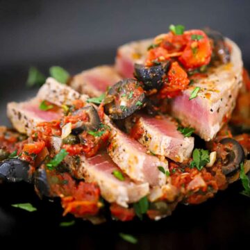 A close-up view of a sliced seared tuna steak that is topped with a Mediterranean sauce of black olives, tomatoes, olives, and garlic.