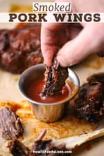 A straight-on view of a person plunging a piece of BBQ pork wings into a small silver vessel filled with homemade barbecue sauce.