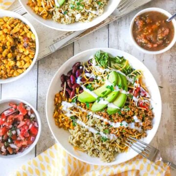 An overhead view of a white bowl holding a serving of turkey burrito bowl topped with slices of avocado, kidney beans, and sour cream and surround by bowls of salsa, pico de galls, and corn.