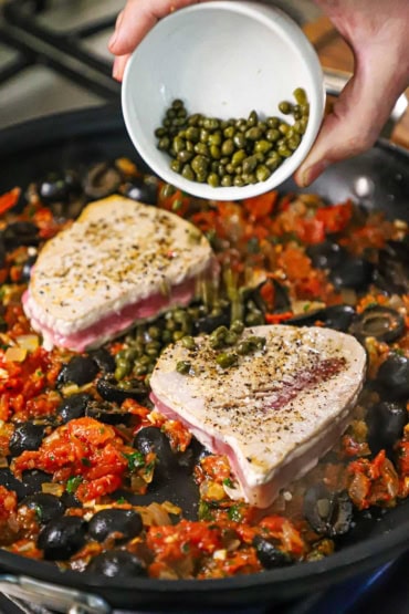 A person dumping a capers from a small white bowl into a large black skillet filled with seared tuna steaks nestled in a tomato and olive sauce.