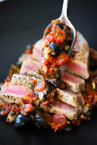 A close-up view of a person spooning a sauce of tomatoes, black olives and capers over sliced Mediterranean tuna steaks.