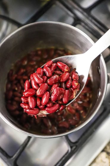 An overhead view of a slotted silver spoon holding a large serving of red kidney beans in a sauce over a saucepan of the simmering beans on a gas stove.
