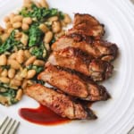 A close-up view of sliced pork tenderloin with a cranberry reduction sauce drizzled on top with a serving of sautéed white beans and spinach next to it.