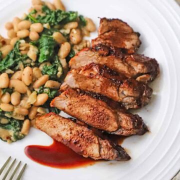 A close-up view of sliced pork tenderloin with a cranberry reduction sauce drizzled on top with a serving of sautéed white beans and spinach next to it.