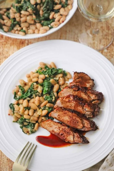 An overhead view of a white dinner plate that is filled with a serving of sliced best-ever pork tenderloin topped with a cranberry reduction and next to a serving of sautéed white beans and spinach.