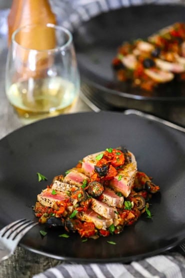 A straight-on view of two black dinner plates side-by-side both filled with Mediterranean tuna steaks with a glass of white wine nearby.