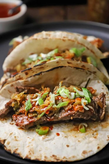 A straight-on view of three Korean beef tacos with kimchi on lightly charred flour tortillas all resting on a black dinner plate.