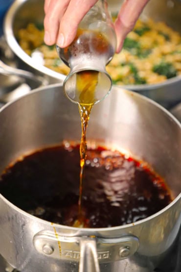 A person pouring Sherry wine vinegar from a small glass carafe into a silver saucepan filled with simmering cranberry juice and molasses.