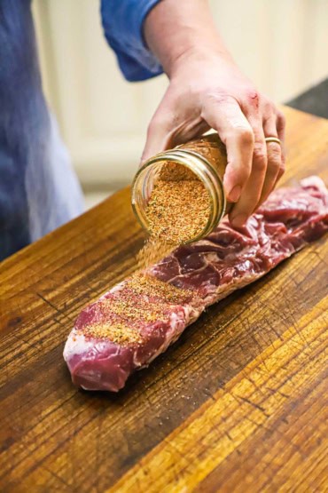 A person dumping pork rub over the top of an uncooked pork tenderloin that is resting on a large wooden cutting board.