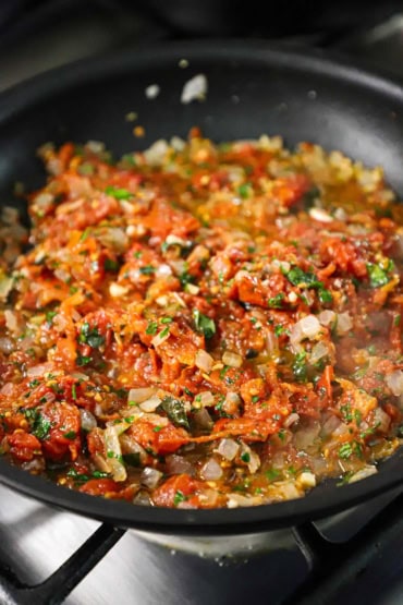 A straight-on view of a large black non-stick skillet filled with a simmering Mediterranean tomato, onion, and black olive sauce.