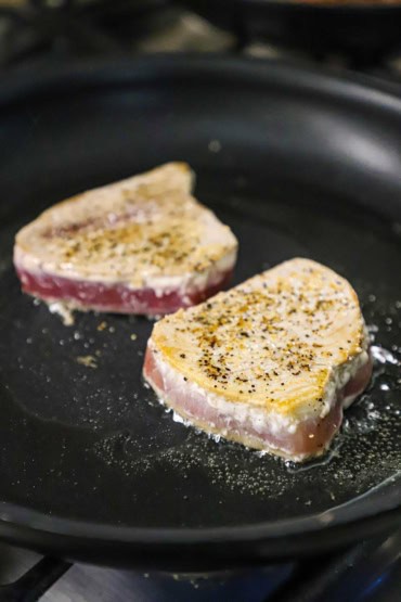 A straight-on view of two medium-sized tuna steaks that have been seasoned with salt and pepper and are being seared in olive oil in a large skillet on the stove.