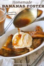 A person using a large silver spoon to drizzle a thin caramel sauce over a serving of bananas foster in small oval dessert dish next to a glass mug of creamed coffee.