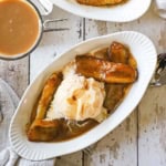 An overhead view of a single serving of bananas foster in an oval white dessert dish with a spoon inserted into the side next to the caramelized bananas and ice cream.