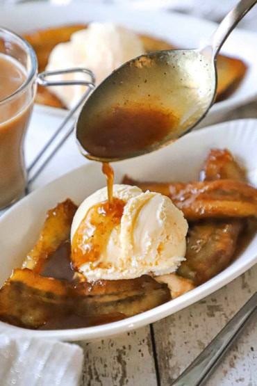 A person using a large silver spoon to drizzle a thin caramel sauce over a serving of bananas foster in small oval dessert dish next to a glass mug of creamed coffee.