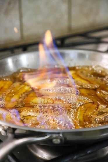 A straight-on view of a silver skillet on gas stove that is filled with simmering banana pieces in a caramel sauce and rum that is has been ignited and is flaming.