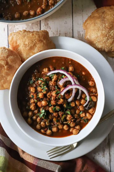 An overhead view of a white bowl sitting on a white plate with a serving of homemade chana masala in the bowl topped with chopped cilantro and onion slivers and several pieces of Indian puff bread nearby.