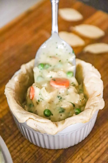 A straight-on view of a large silver spoon being used to transfer the filling of a chicken pot pie into a large ramekin that has been lined with a pie dough.