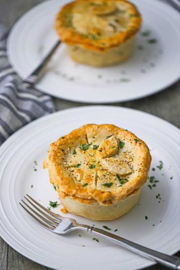 Two unbroken chicken pot pies that both have a top and bottom crust and are sitting on white dinner plates and garnish with finely chopped parsley.