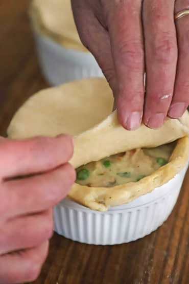 A person placing a piece of uncooked pie dough on top of a large ramekin that has been lined with a par-baked pie dough and filled with chicken pot pie filling.