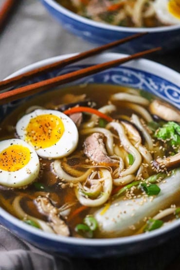 A straight-on view of a large shallow colorful bowl filled with udon noodle soup with pork including two halves of hard boiled eggs and Chinese cabbage in the soup.