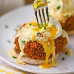 A close-up view of a fork being used to break open a poached egg that is resting on top of a crab cake that is on the bottom of a toasted English muffin.