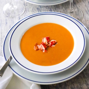 A close-up view of a bowl of lobster bisque in a shallow white bowl with a blue thin-line rim and there is fresh lobster chunks resting on top.