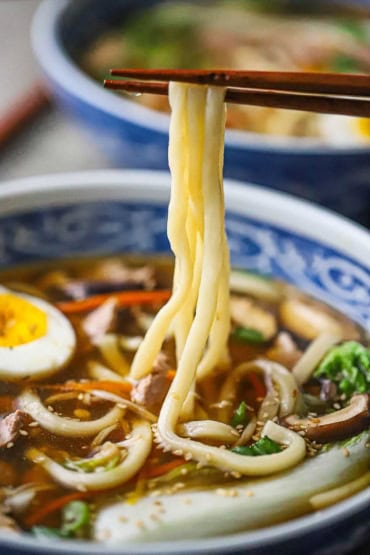 A close-up view of a pari of chop sticks being used to lift up cooked udon noodles from a bowl of udon soup with pork.