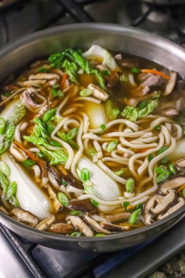 A view of a large stainless steel skillet filled with udon soup with pork with several cooked Chinese cabbage leaves and sliced mushrooms visible within the soup.