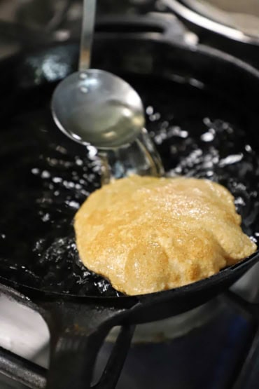 A close-up view of an Indian puff bread called poori that is being deep-fried in a black cast-iron skillet filled with hot oil and a small ladle being used to pour the hot oil over the top of the poori.