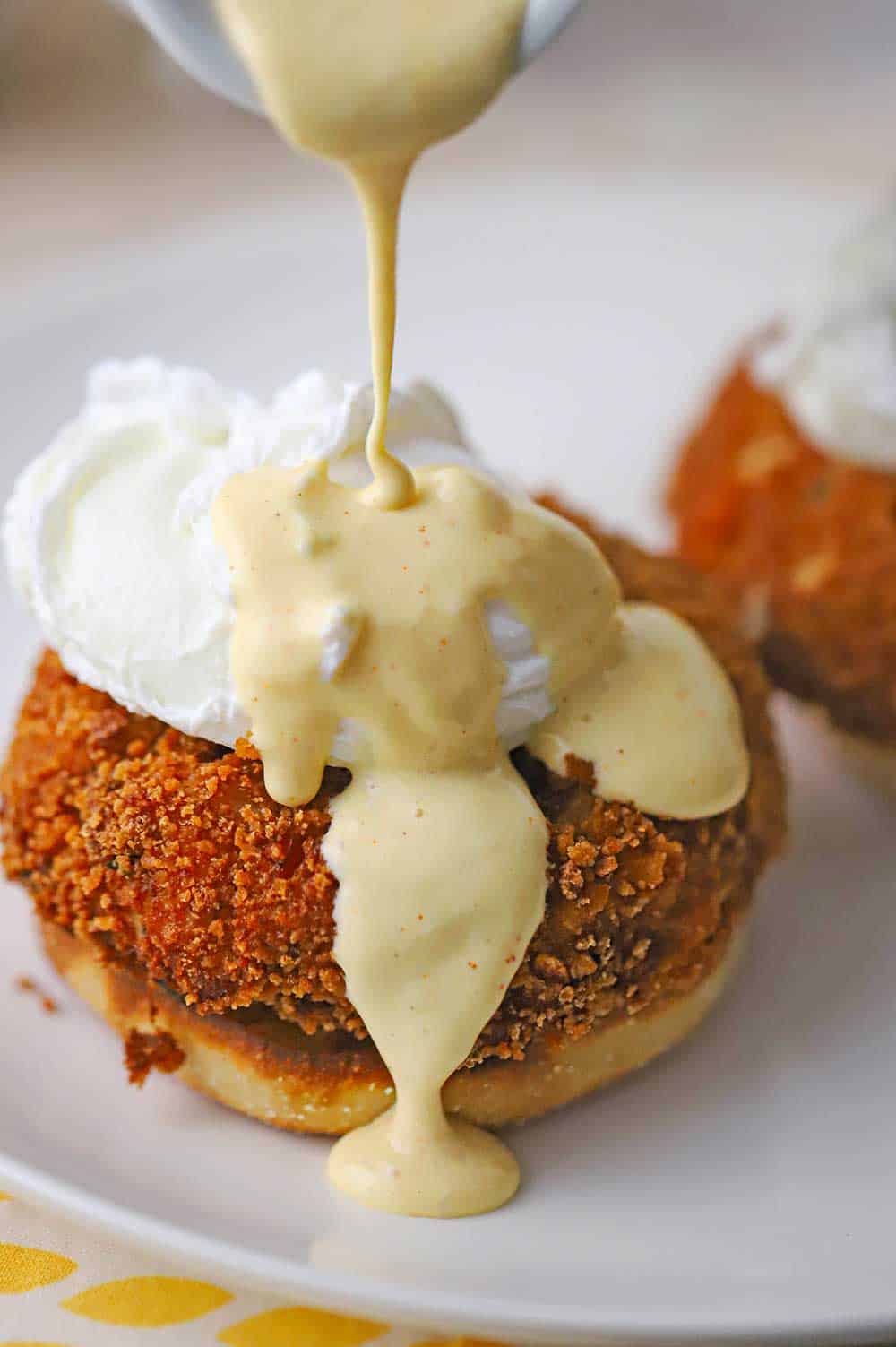 A close-up view of hollandaise sauce being poured from a small white pitcher onto a poached egg that is resting on top of a crispy crab cake and the bottom of an English muffin.